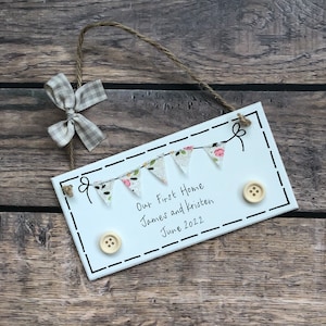 May include: White wooden sign with a string for hanging, decorated with fabric bunting and buttons. The sign reads "Our First Home James and Kristen June 2022".