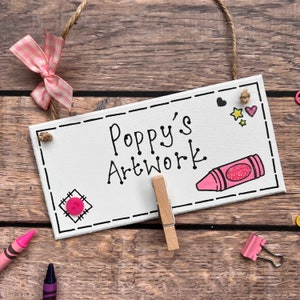 May include: White wooden sign with black text that reads "Poppy's Artwork". The sign has a pink button, a pink crayon, and pink hearts and stars drawn on it. A wooden clothespin is attached to the sign.