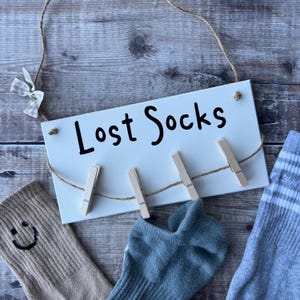 May include: White wooden sign with the text "Lost Socks" and a string with clothespins hanging from it. There are three socks hanging on the clothespins, one brown, one blue, and one striped.