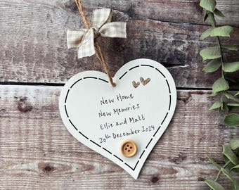 May include: White wooden heart-shaped sign with the text "New Home New Memories Ellie and Matt 20th December 2024". The sign has a brown button and two small gold glitter hearts.