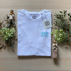 May include: A white t-shirt with the word "DAD" embroidered in light blue, along with "est. 2021". The shirt has a tag attached and a small logo on the collar. Decorative greenery and figurines surround the shirt.