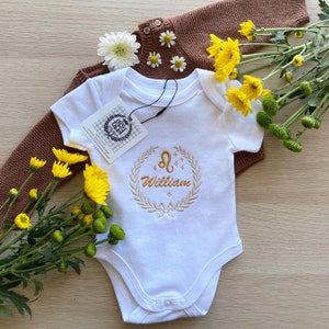 Op de afbeelding: Witte babybody met gouden borduursel van de naam "William" in een lauwerkrans, met een Leeuw-dierenriemteken. Een bruine gebreide trui bevindt zich achter de body. Gele bloemen en madeliefjes zijn rond de kleding gerangschikt.