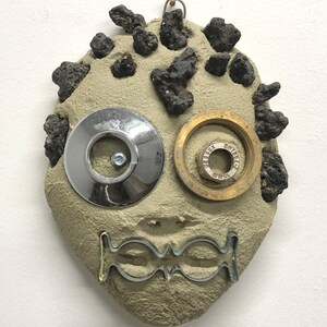 Op de afbeelding: Een handgemaakte wanddecoratie in de vorm van een gezicht. Het heeft een lichtbruine, getextureerde basis met donkere, ruwe uitsteeksels. De ogen zijn gemaakt van een chromen schijf en een messing ring. De mond is een zilverkleurig, gestileerd ontwerp.