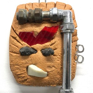 Könnte beinhalten: Eine Wandskulptur in Mischtechnik mit einer beigefarbenen, strukturierten Basis, die einen roten Glassplitter, schwarze Steine und ein weißes Stück aufweist. Metallrohre und -fittings sind angebracht, zusammen mit zwei Laschen zum Aufhängen.