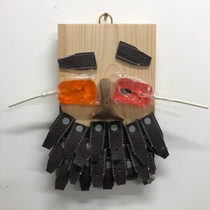 May include: A quirky artwork depicting a face on a wooden board. Dark brown leather pieces form the eyes, a wooden carving is the nose, and dark brown leather straps make up the mouth. Orange and red lollipops are used for the cheeks.