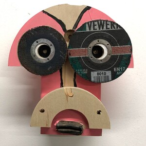 Op de afbeelding: Abstracte sculptuur in de vorm van een gezicht. Het heeft twee cirkelvormige slijpschijven als ogen, een roze geschilderde hoofdvorm en een houten boogvormige mond. Een uniek kunstwerk.