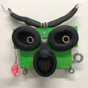 Könnte beinhalten: Ein skurriles Kunstwerk, das einem Gesicht ähnelt und aus recycelten Kopfhörern und Hardware besteht. Es zeigt schwarze Ohrmuscheln, eine grüne Holzbasis und Metalldetails. Enthält einen herzförmigen rosa Anhänger und andere hängende Elemente.