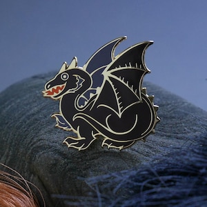 Puede incluir: Un pin de esmalte de dragón negro con ribete dorado, que presenta un diseño detallado de una criatura mítica con alas y boca roja. El pin está sobre una superficie gris texturizada con hilo naranja y azul en el fondo.