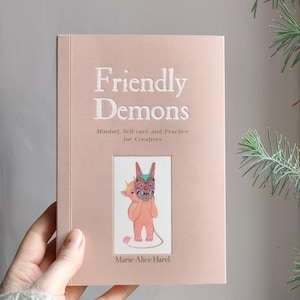 Peut inclure: Un livre à couverture rigide rose clair intitulé "Friendly Demons" avec le sous-titre "Mindset, Self-care and Practice for Creatives" par Marie-Alice Harel. La couverture présente un texte en relief blanc et une illustration colorée de deux démons de dessin animé.