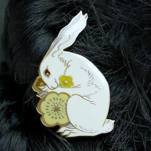 Könnte beinhalten: Ein weißer Emaille-Kaninchen-Anstecker mit goldenen Verzierungen, das eine gelbe Blume hält. Das Kaninchen hat detaillierte geätzte Linien und ein rotes Auge. Der Anstecker ist vor einem dunklen Hintergrund platziert.