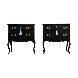 May include: Two black painted wooden bedside tables with ornate carved legs and gold drawer pulls. Each bedside table has two drawers with a tassel pull on the top drawer.