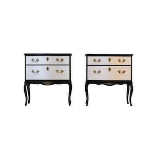 May include: Two black and white bedside tables with two drawers each. The drawers have gold handles and the tables have gold accents on the legs.