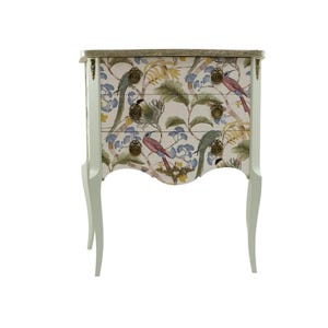 Può includere: Una cassettiera bianca dipinta con due cassetti. I cassetti sono decorati con una stampa floreale con uccelli e foglie. La cassettiera ha gambe ornate e ferramenta dorata.