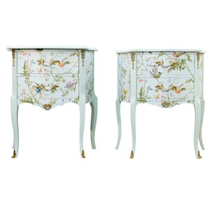 May include: A pair of matching bedside cabinets in a pale blue hue, featuring a floral and bird design. Each cabinet has two drawers and gold detailing. The curved legs have gold-coloured feet. (Measurements in cm)