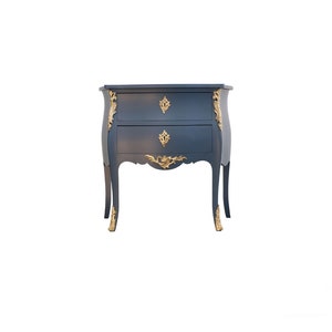 Può includere: Un comodino a due cassetti blu scuro con dettagli dorati e decorazioni elaborate. Il comodino ha un fronte e gambe curve.