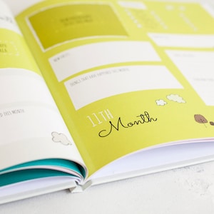 K&ouml;nnte beinhalten: Ein gelbes und wei&szlig;es Baby-Erinnerungsbuch mit dem Text "11th Month" in Schwarz. Das Buch hat einen Bereich f&uuml;r Fotos, einen Bereich f&uuml;r neue Errungenschaften und einen Bereich f&uuml;r Dinge, die in diesem Monat passiert sind.
