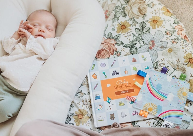 K&ouml;nnte beinhalten: Ein Baby schl&auml;ft in einem wei&szlig;en, nistf&ouml;rmigen Kissen auf einem floral gemusterten Stoff. Ein blaues und oranges Baby-Erinnerungsbuch mit bunten Illustrationen liegt auf dem Stoff, zusammen mit einem blauen und gelben Stoffquadrat mit einem Regenbogen- und Sonnenmotiv.