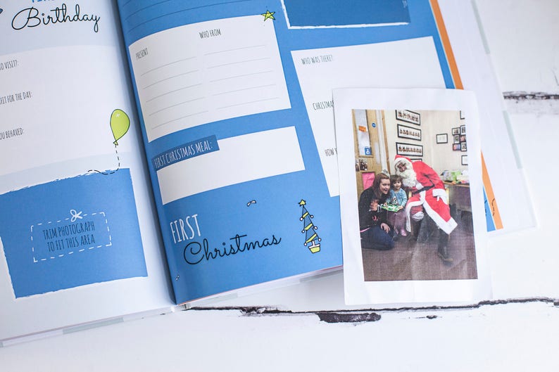 Puede incluir: Un libro de recuerdos para beb&eacute;s azul y blanco con una foto de un ni&ntilde;o con Pap&aacute; Noel. El libro tiene las palabras "Primera Navidad" en la portada.