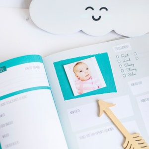 Puede incluir: Un libro de recuerdos para beb&eacute;s con una cubierta azul turquesa y una foto de una ni&ntilde;a peque&ntilde;a en la p&aacute;gina. La p&aacute;gina tiene secciones para registrar las primeras palabras del beb&eacute;, c&oacute;mo se mueve el beb&eacute;, su temperamento y sus nuevos dientes.