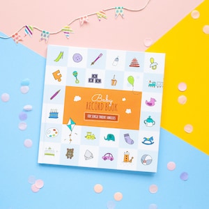 Puede incluir: Un libro de recuerdos para bebés de color blanco y naranja con el título "Baby Record Book" y el texto "Para familias monoparentales". La portada presenta una variedad de ilustraciones coloridas de artículos para bebés, que incluyen un sonajero, un tren de juguete, un elefante de peluche y un chupete.