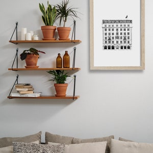 Puede incluir: Tres estantes de madera con soportes de metal negro est&aacute;n montados en una pared blanca. Los estantes est&aacute;n decorados con plantas en macetas, libros y botellas de vidrio. Un dibujo en blanco y negro de un edificio enmarcado cuelga de la pared sobre los estantes.
