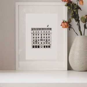 Puede incluir: Un dibujo en blanco y negro de un edificio con una bandera en la parte superior, enmarcado en un marco blanco y colocado en un estante blanco. El estante est&aacute; decorado con un jarr&oacute;n de flores secas y una pila de libros.