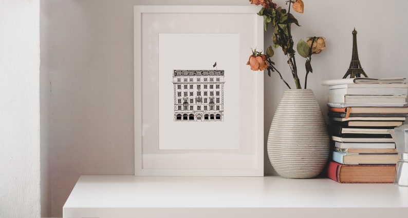 Puede incluir: Un dibujo en blanco y negro de un edificio con una bandera en la parte superior, enmarcado en un marco blanco y colocado en un estante blanco. El estante est&aacute; decorado con un jarr&oacute;n de flores secas y una pila de libros.