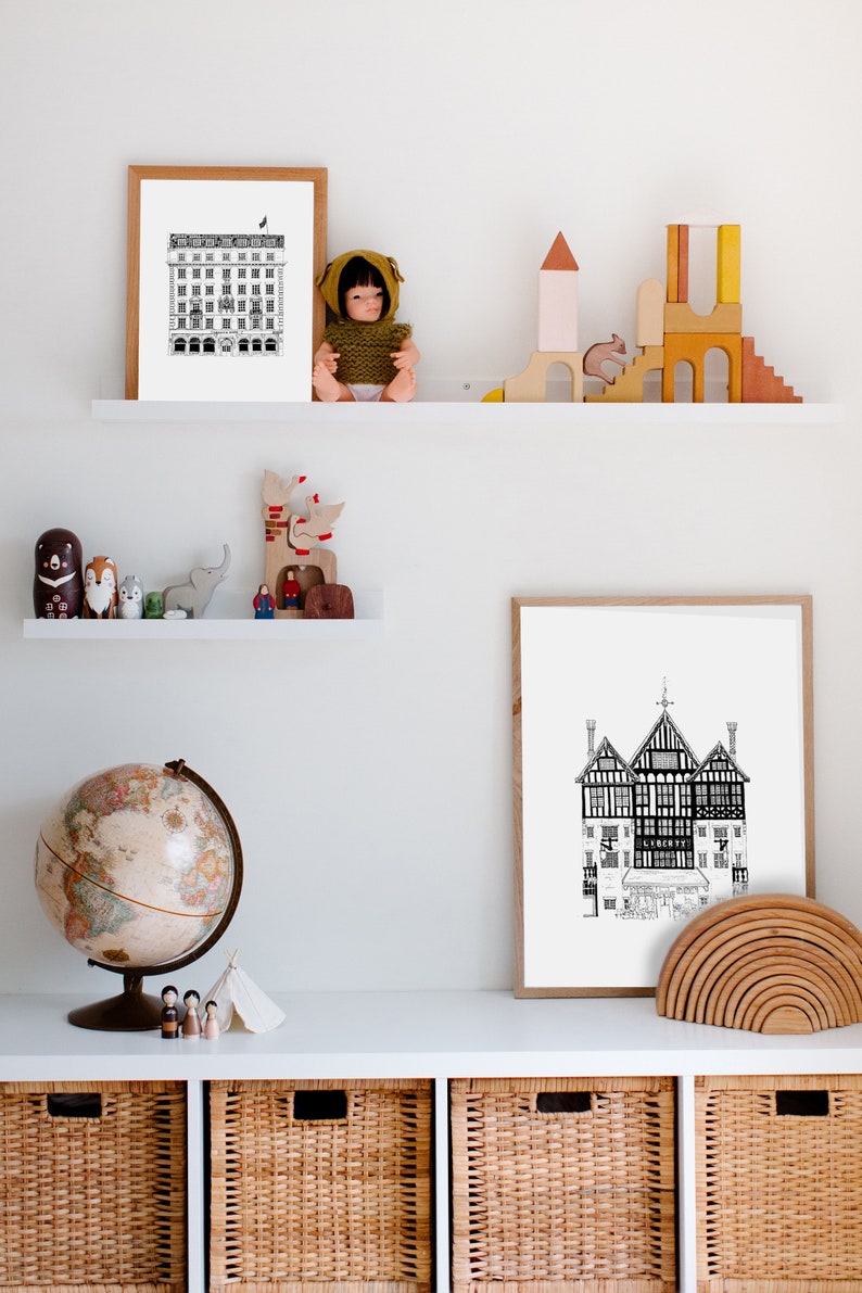 Puede incluir: Un estante blanco con dos ilustraciones en blanco y negro enmarcadas de edificios, un globo terr&aacute;queo y un juego de bloques de construcci&oacute;n de madera. El estante est&aacute; sobre un conjunto de tres cestas de mimbre.