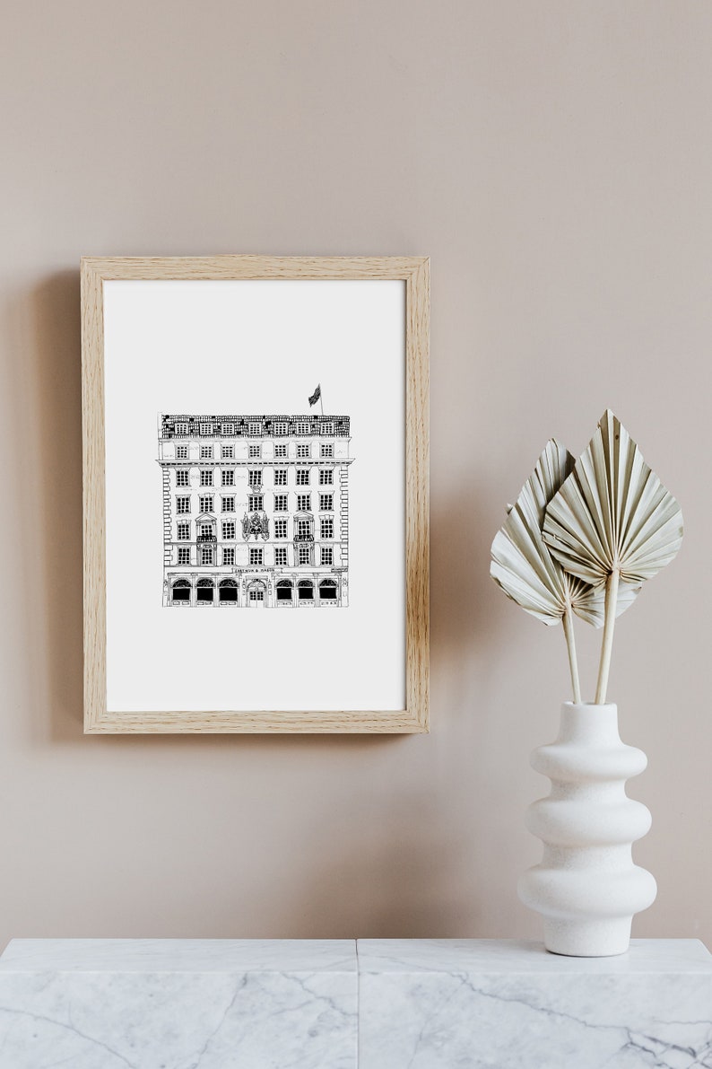 Puede incluir: Un dibujo en blanco y negro de un edificio con una bandera en la parte superior. El dibujo est&aacute; enmarcado en un marco de madera claro y est&aacute; colgado en una pared de color beige claro. Un jarr&oacute;n blanco con hojas de palma secas est&aacute; sobre una superficie de m&aacute;rmol blanco.