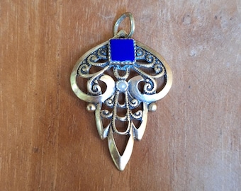 May include: A gold pendant with a blue enamel square and intricate filigree design. The pendant is on a wooden surface.