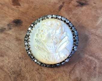 May include: A white cameo brooch with a detailed carving of a woman's face surrounded by a silver-toned setting with rhinestones.