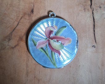 Peut inclure: Un poudrier rond argenté avec un motif floral. Le poudrier présente un fond bleu clair avec un motif radial et une orchidée peinte dans des tons de rose, vert et jaune. Une petite boucle est en haut.