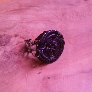 May include: A black ring with a large, round, ornate, black button-like design. The ring is made of metal with a decorative, openwork design.