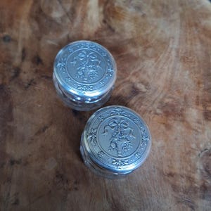 May include: Two small, silver-colored, round containers with ornate floral and ribbon designs on the lids. The containers are on a wooden surface with a natural wood grain pattern.
