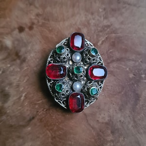 Puede incluir: Un broche de plata con piedras de vidrio rojo y verde engastadas en un diseño de filigrana.