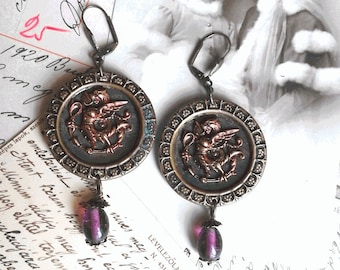 May include: A pair of earrings with a bronze-coloured circular pendant featuring a detailed embossed design of a mythical creature. Each pendant is accented with a dark purple glass bead.