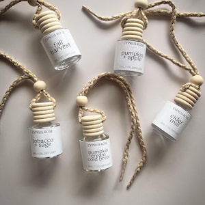 Op de afbeelding: Vier kleine glazen flesjes met houten dopjes en beige touw hangen aan een wit oppervlak. Elk flesje heeft een wit etiket met de tekst "CYPRUS ROSE" en een andere geurbeschrijving: "fall harvest", "tobacco + sage", "pumpkin + apple" en "pumpkin cream cold brew".