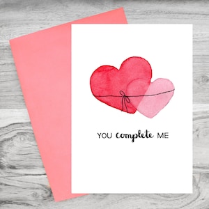 May include: A white card with two watercolor hearts, one red and one pink, tied together with a string. The text "YOU complete ME" is printed below the hearts.