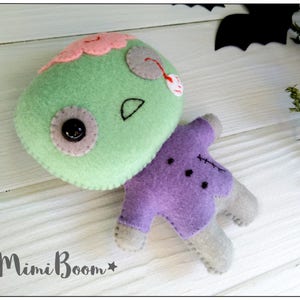 May include: A handmade felt zombie doll with a green head, pink brain, and purple body. The doll has a button eye and stitched details. The Mimi Boom logo is visible in the bottom left corner. Perfect for Halloween.