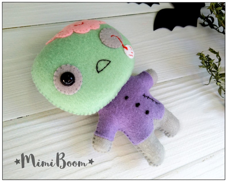 May include: A handmade felt zombie doll with a green head, pink brain, and purple body. The doll has a button eye and stitched details. The Mimi Boom logo is visible in the bottom left corner. Perfect for Halloween.