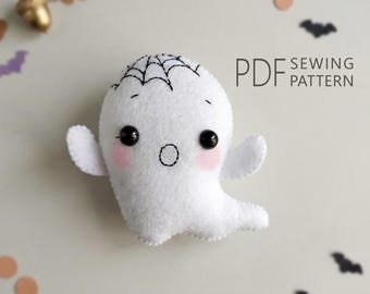May include: A white felt ghost plushie featuring black embroidered eyes, a small circular mouth, and a spiderweb design on its head. The ghost has soft pink cheeks and outstretched arms. The text PDF SEWING PATTERN is printed to the right.