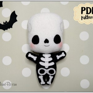 May include: A white felt skeleton plush toy with black felt details. The toy has a smiling face with black eyes and a black mouth. The toy is wearing a black felt skeleton costume. The toy is sitting on a white and gray polka dot background. The text "PDF pattern" is in a brown cloud shape in the upper right corner of the image.