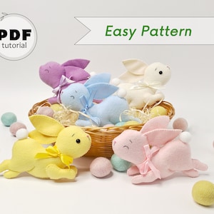 May include: Five felt Easter bunny plush toys in various colours, including pink, yellow, blue, white, and purple. The bunnies are sitting in a wicker basket with pastel coloured pom poms scattered around them. The text "Easy Pattern" is displayed above the bunnies.