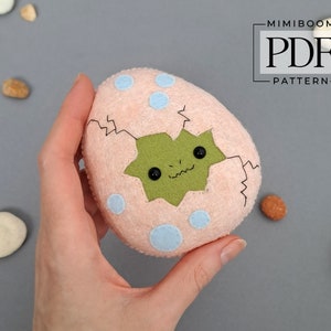 May include: A plush, peach-colored egg with light blue polka dots and a cracked opening revealing a green dinosaur. The dinosaur has black button eyes and a stitched smile. The text "MIMIBOOM PDF PATTERN" is in the upper right corner.