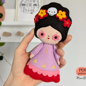May include: A handmade felt doll with black hair, decorated with yellow and red flowers and a skull. The doll is dressed in a lavender dress with yellow embroidered details and a red skirt. The doll is held in a hand.