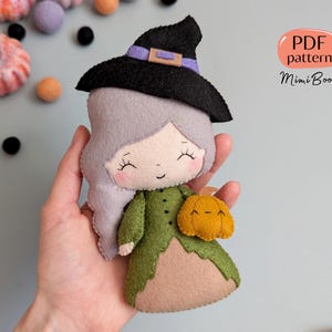 Puede incluir: Muñeca de bruja de fieltro hecha a mano con sombrero negro, vestido verde y una pequeña calabaza naranja. La muñeca tiene pelo gris y una cara sonriente. La imagen incluye el texto "PDF pattern MimiBoom".