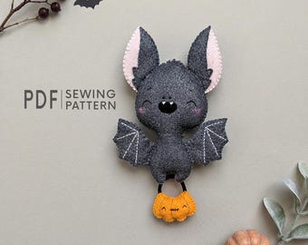 May include: A gray felt bat plush toy with pink inner ears, white stitched wing details, and a black nose. The bat holds a small orange felt pumpkin with a stitched face. The text PDF SEWING PATTERN is printed to the left of the bat.