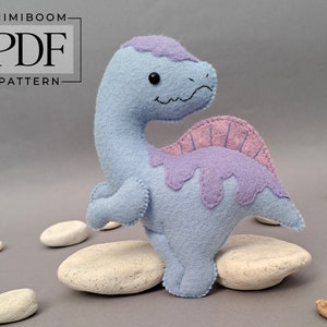Op de afbeelding: Een lichtblauwe vilten dinosaurus met een roze en paarse vilten rug. De dinosaurus staat op een witte steen.