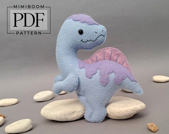 May include: A light blue felt dinosaur with a pink and purple felt back. The dinosaur is standing on a white rock.