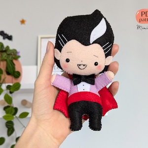 May include: A handmade felt vampire doll with black hair, a white streak, and a black bow tie. The doll wears a pink shirt, black trousers, and a red cape. The doll has a smiling face and the text "PDF pattern MimiBoom".